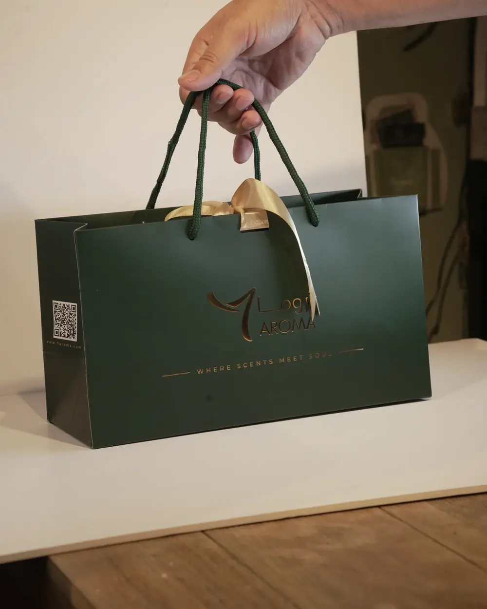 Photograph: a hand holding the deep green 7 AROMA gift bag with a gold-foil logo and a wide champagne satin bow
